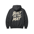 A dark gray hoodie shown from the back, featuring a large cream-colored graphic text print that reads 'BUILT ON THE MAT' in a stylized brushstroke font.